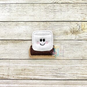 May include: A felt applique of a smiling marshmallow on a graham cracker with chocolate. The marshmallow is white with pink cheeks and a black smile.