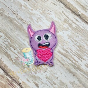 May include: Purple felt monster with a pink heart in its hands. The monster has a big smile and two large eyes.