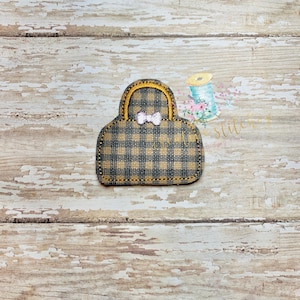 Op de afbeelding: Een klein zwart en bruin geruit handtasje met een gele handvat en een witte strik. Het handtasje ligt op een houten achtergrond.