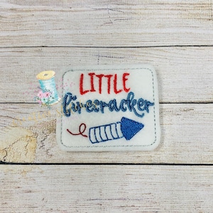 May include: White felt patch with red and blue embroidery. The text "LITTLE firecracker" is embroidered in red and blue. A blue and white firework is embroidered below the text.