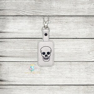 May include: A white leather key chain with a silver key ring and a black embroidered skull design.