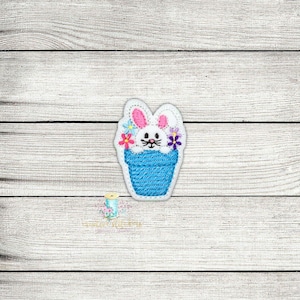 May include: A white bunny with pink ears and a blue flower pot with pink, purple, and blue flowers. The bunny is peeking out of the pot.