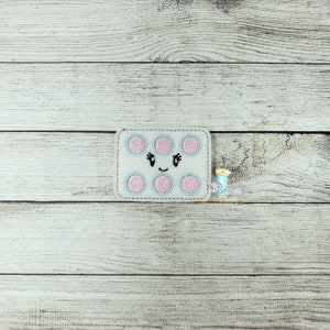 May include: A rectangular, white felt patch with a smiling face and six pink circles outlined in light blue. The patch has a black stitched mouth and eyes. The background is a light wood grain.