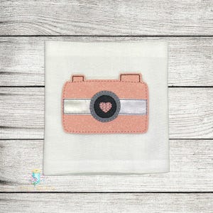 Puede incluir: Una cámara de fieltro rosa con una franja plateada y un pequeño corazón rosa en el centro del objetivo. La cámara está sobre un fondo blanco.