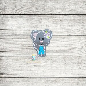 Peut inclure: Souris grise brodée portant une salopette bleue et une fleur violette. La souris a un nez et des oreilles roses, avec des coutures blanches sur les bords. Le fond est un grain de bois clair.