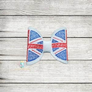 May include: A white bow tie featuring the Union Jack flag design. The bow tie has a white center and the flag design is in red, white, and blue. The bow tie is likely made of fabric and is set against a light wood grain background.