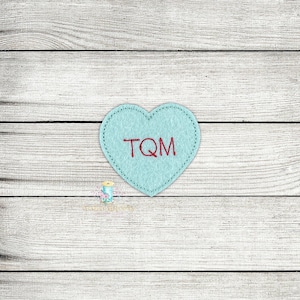 May include: A light blue felt heart with the letters "TQM" embroidered in red thread.