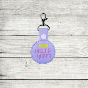Puede incluir: Llavero redondo morado con una corona dorada y el texto "drama queen" en letras rosas.
