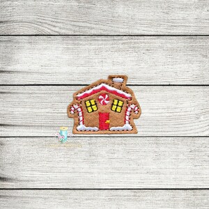 Op de afbeelding: Een gingerbread house applique met witte frosting, rode snoepstokken en gele ramen. Het huis is bruin en heeft een rode deur.