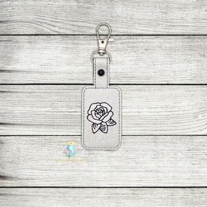 May include: A white leather keychain with a black embroidered rose design. The keychain has a silver metal key ring.