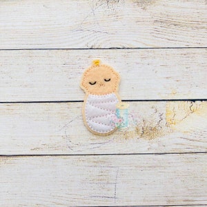 Puede incluir: Una aplicaci&oacute;n de fieltro de un beb&eacute; envuelto en una manta rosa con una cinta amarilla. El beb&eacute; tiene los ojos cerrados y una sonrisa cosida.