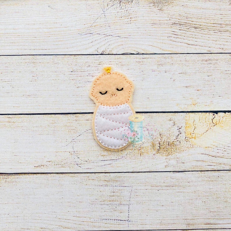 Puede incluir: Una aplicaci&oacute;n de fieltro de un beb&eacute; envuelto en una manta rosa con una cinta amarilla. El beb&eacute; tiene los ojos cerrados y una sonrisa cosida.
