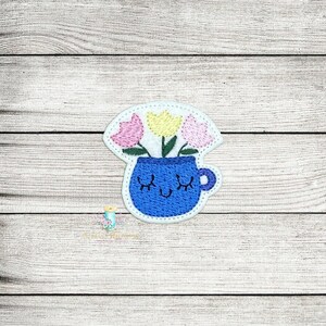 Puede incluir: Una taza azul con una cara sonriente y los ojos cerrados, sosteniendo tres flores: rosa, amarilla y rosa. Las flores están en una maceta blanca.