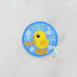 Könnte beinhalten: Ein gestickter Aufnäher mit einer Gummi-Ente und dem Text "Rub-a-Dub-Dub" in Blau und Gelb.