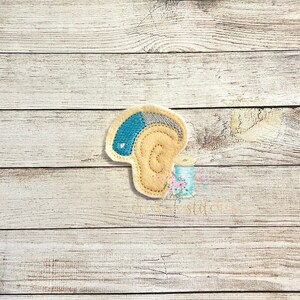 Puede incluir: Una aplicación de fieltro de una oreja azul claro y gris con un corazón blanco en el centro. La oreja es beige y tiene un patrón de espiral.