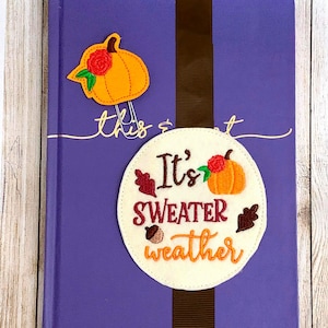 Può includere: Un segnalibro in feltro bianco con il testo "It's sweater weather" ricamato in marrone e rosso. Il segnalibro presenta una piccola zucca ricamata e foglie. Un nastro marrone è attaccato al segnalibro.