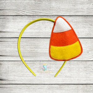 Puede incluir: Una diadema amarilla con un caramelo de maíz de fieltro adjunto. El caramelo de maíz es naranja, amarillo y blanco con un diseño cosido.