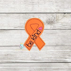 Pode incluir: Uma fita laranja com as palavras "be kind" bordadas em preto. Um pequeno coração rosa é costurado perto do topo. A fita está em um fundo de madeira branca.