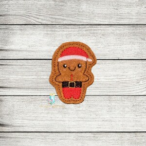 Puede incluir: Un hombre de jengibre con un gorro de Papá Noel rojo y blanco con un cinturón negro. El hombre de jengibre sonríe y tiene la cara marrón.