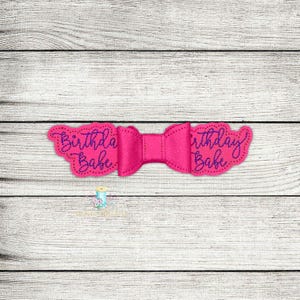 Puede incluir: Un lazo rosa con un centro blanco y las palabras "Birthday Babe" bordadas en morado a cada lado.