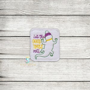 May include: A white square patch with a green outline featuring a cartoon alligator wearing a purple hat and yellow collar. The text "Let the GOOD TIMES roll" is embroidered in purple and yellow.