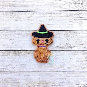 Può includere: Un gatto di glitter marrone che indossa un cappello da strega nero con un bordo verde. Il gatto ha un collare viola e guance rosa.