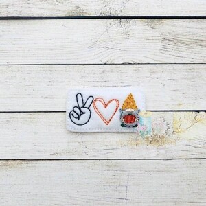 May include: White felt patch with a peace sign, a red heart, and a gnome with a red hat and a pumpkin in its hands. The text "Peace Love Gnome" is embroidered on the patch.