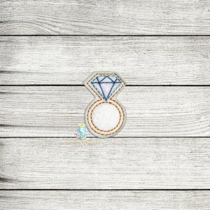 Puede incluir: Un anillo de fieltro blanco con un diamante azul en la parte superior. El anillo está bordeado con hilo dorado.