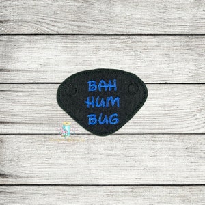 Peut inclure: Un cache-œil en feutre noir avec une broderie bleue qui dit "Bah Hum Bug".