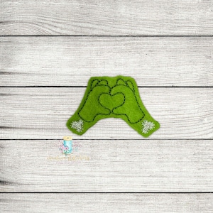 Op de afbeelding: Groene vilt hand applicatie met een hartvorm die door de vingers wordt gevormd. Het hart is omlijnd met zwarte draad en de handen hebben witte kralen voor detail.