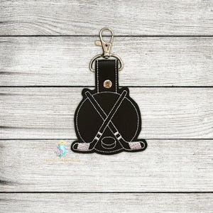 Può includere: Portachiavi nero a tema hockey con chiusura argentata. Il portachiavi presenta un design circolare con bastoni da hockey incrociati e un disco, il tutto delineato da cuciture bianche. Il design è attaccato a un cinturino in pelle nera.