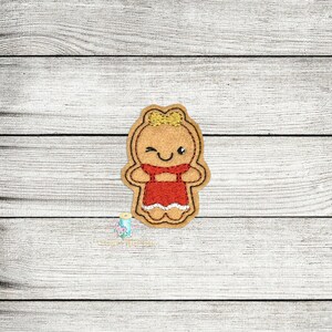 May include: A gingerbread person with a red dress and a yellow bow in her hair. She is winking and smiling.