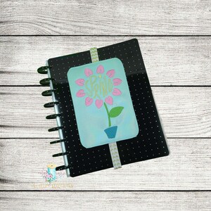 May include: A black notebook with white polka dots and a green spiral binding. A light blue bookmark with a flower design and the word "SPRING" in yellow is on top of the notebook. The bookmark has a green and white patterned ribbon.