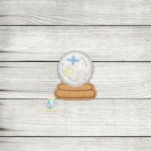 May include: A white felt snow globe with a blue star and a yellow crescent moon inside. The snow globe is sitting on a brown felt base.