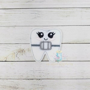May include: White felt tooth with a happy face, eyelashes, and a gray felt brace. The tooth is on a white wood background.