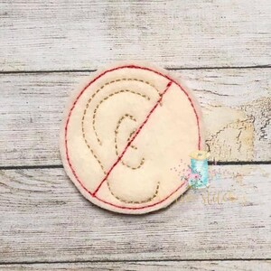 Puede incluir: Un círculo de fieltro blanco con un contorno cosido rojo. Dentro del círculo hay una imagen de una oreja cosida en oro con una línea roja que la atraviesa.