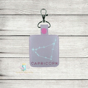 Puede incluir: Un llavero de cuero morado con un anillo de llave plateado y un bot&oacute;n rosa. El llavero presenta una constelaci&oacute;n bordada en azul claro con la palabra "CAPRICORN" en letras marrones.