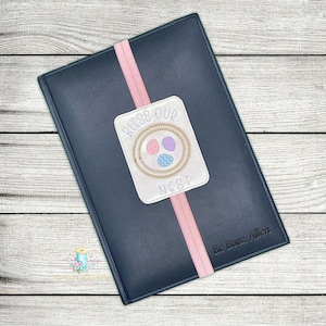May include: A black leather journal with a pink bookmark. The bookmark has an embroidered design that says "Bless Our Nest" with a nest and three eggs.
