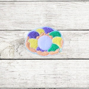 May include: An embroidered patch shaped like a king cake, featuring purple, green and yellow colours. The cake has a white centre.