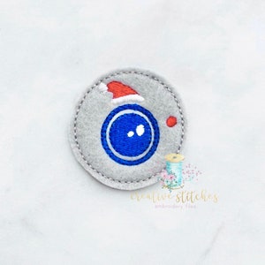 May include: A grey felt circle with a blue button and a red Santa hat on top. The button has two white dots for eyes.