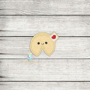 May include: A light brown fortune cookie with a smiling face and a red heart on a white envelope. The cookie is on a light wood background.