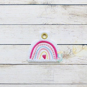 May include: A white felt rainbow keychain with a pink, blue, and yellow rainbow and a small pink heart in the center. The keychain has a gold metal ring.