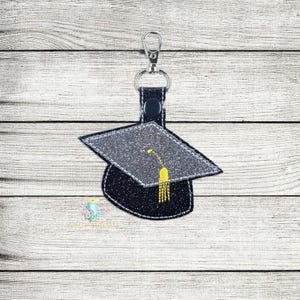 May include: A black graduation cap keychain with a silver key ring and a yellow tassel. The cap is made of felt and has a glitter finish.
