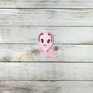 May include: Pink felt stethoscope with a smiling face and black eyes. The stethoscope has a red top and a black cord.