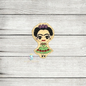 May include: Embroidered felt patch of Frida Kahlo, a famous Mexican artist, wearing a green dress with red flowers and a purple flower crown.