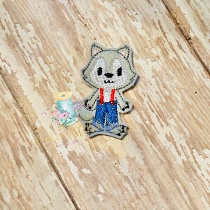 Peut inclure: Un loup brodé gris portant une salopette bleue et des bretelles rouges. Le loup a un visage blanc et des yeux noirs.