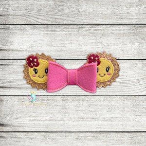May include: A pink bow with two yellow sun-shaped embellishments with smiling faces and red flowers. The sun embellishments have a gold glitter finish.