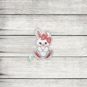 Könnte beinhalten: Ein weißer gestickter Hase mit rosa Ohren und einer rosa Schleife. Der Hase sitzt mit seinen Pfoten vor sich.
