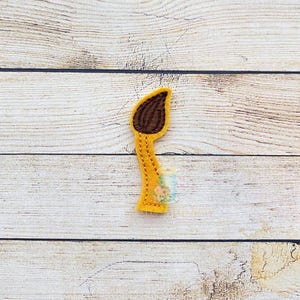 May include: A felt applique in the shape of a paintbrush. The paintbrush has a brown brush head and a yellow handle. The handle is decorated with small yellow stitches. The background is a light wood grain.