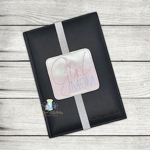 May include: A black leather journal with a silver bookmark and a white patch that says "Girl Mom" in pink and blue lettering. The journal has the name "Be Booz Allen" embossed on the bottom right corner.